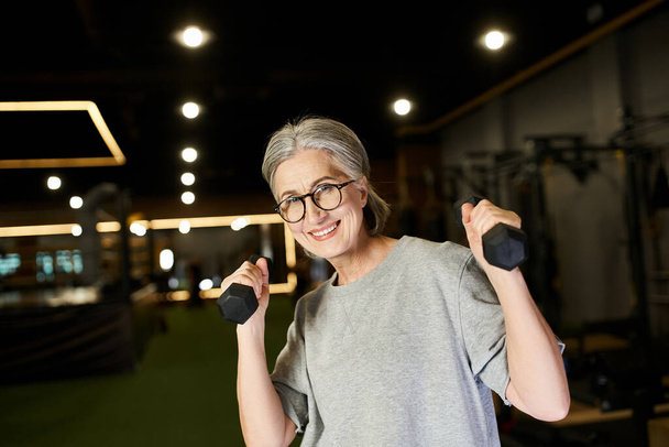 mujer madura alegre con gafas y canas haciendo ejercicio con mancuernas y sonriendo a la cámara - Foto, Imagen