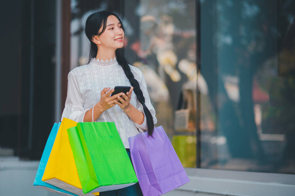 Una mujer sostiene bolsas de la compra y mira su teléfono. Ella sonríe y disfruta de su experiencia de compra - Foto, Imagen