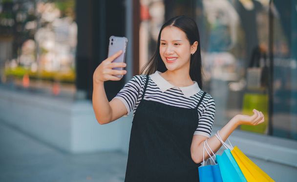 Une femme prend un selfie avec son téléphone portable tout en tenant des sacs à provisions. Elle sourit et profite de son expérience d'achat - Photo, image
