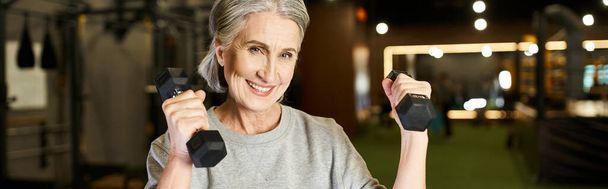 pretty jolly mature woman in sportswear training with dumbbells and smiling at camera, banner - Photo, Image