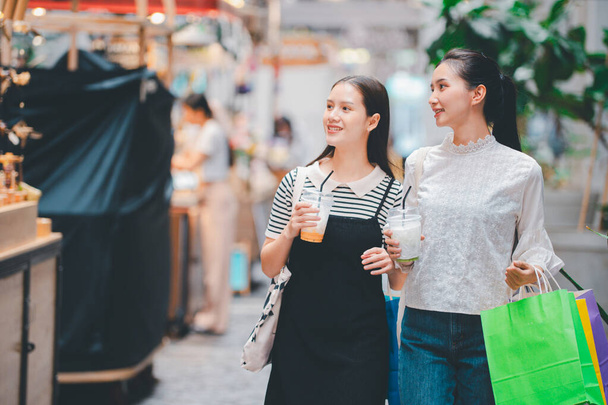 Two women are walking down a street, one of them holding a green bag. They are both smiling and holding cups - Photo, Image
