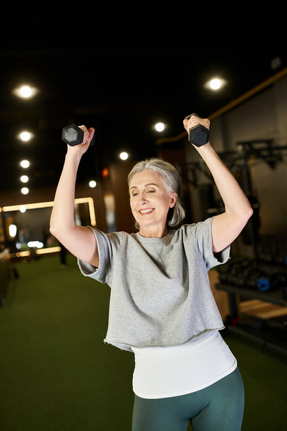 gut aussehende lächelnde reife Frau in bequemer Sportbekleidung, die glücklich mit Hanteln im Fitnessstudio trainiert - Foto, Bild