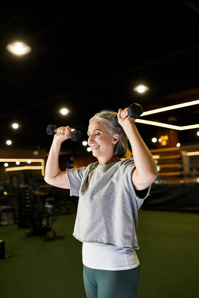 mujer mayor deportivo en acogedor entrenamiento de atuendo con pesas y sonriendo felizmente mientras está en el gimnasio - Foto, imagen