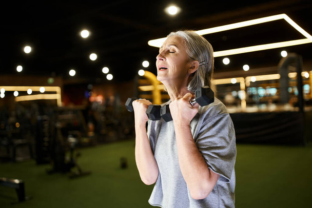 sportowiec wesoły dojrzała kobieta z siwymi włosami w odzieży sportowej szkolenia z hantlami podczas gdy w siłowni - Zdjęcie, obraz