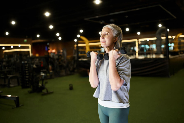 athletic appealing mature sportswoman in comfy attire exercising hard with dumbbells while in gym - Photo, Image