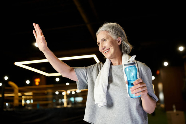 красивая, веселая старшая спортсменка с полотенцем на плечах и активно машущей бутылкой воды - Фото, изображение