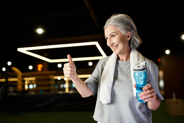 merry athletic mature woman in sportswear with towel on shoulders with water bottle showing thumb up - Photo, Image