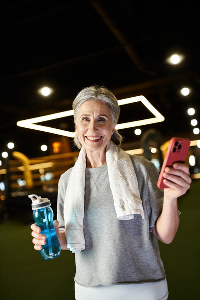 alegre mujer mayor con toalla en los hombros sosteniendo teléfono y botella y sonriendo a la cámara - Foto, imagen