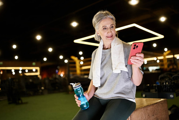 fröhliche reife grauhaarige Sportlerin in bequemer Kleidung, die mit einer Wasserflasche in der Hand auf das Telefon blickt - Foto, Bild