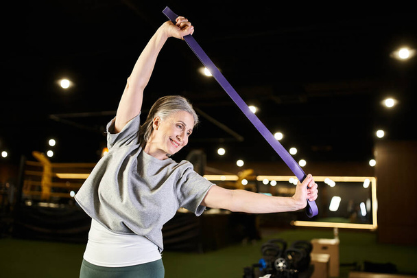 fröhliche reife Sportlerin in bequemer Kleidung, die im Fitnessstudio aktiv mit Fitness-Expander trainiert - Foto, Bild