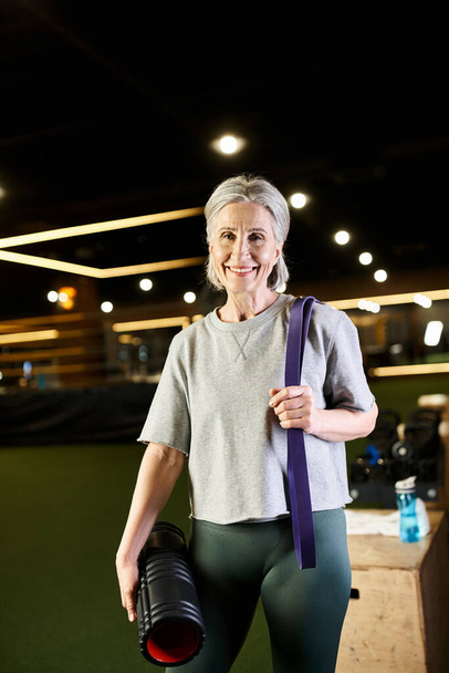 весёлая взрослая спортсменка позирует с экспандером для фитнеса и мешком для веса и улыбается в камеру - Фото, изображение