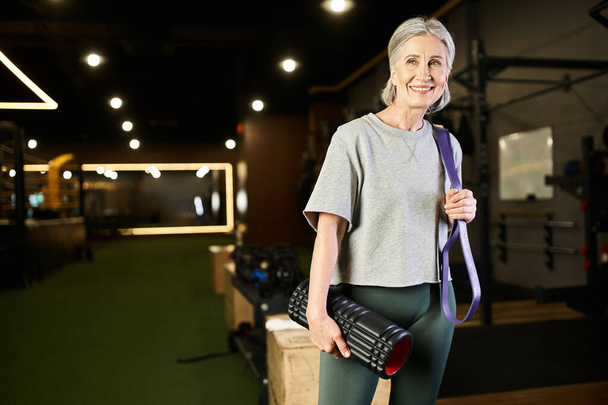 atractiva mujer mayor alegre posando en el gimnasio con expansor de fitness y bolsa de peso y mirando hacia otro lado - Foto, imagen