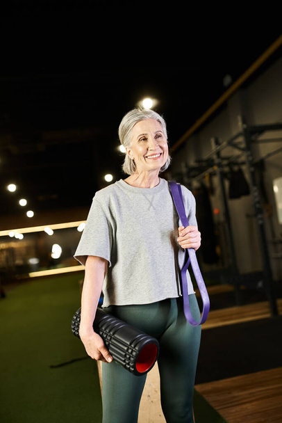alegre atractiva mujer madura posando en el gimnasio con expansor de fitness y bolsa de peso y mirando hacia otro lado - Foto, Imagen
