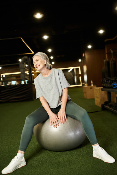 fröhliche reife Frau in Sportbekleidung posiert mit Fitness-Expander und Gewichtsbeutel und lächelt in die Kamera - Foto, Bild