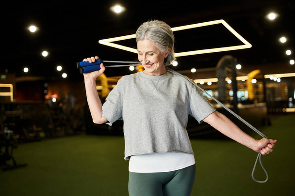 joyeuse mature belle femme en vêtements de sport posant avec corde à sauter dans la salle de gym et regardant loin - Photo, image