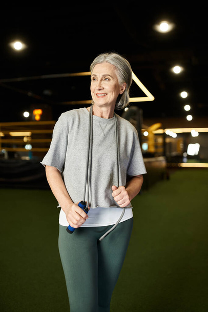 joyful senior appealing woman in sportswear posing with skipping rope in gym and looking away - Photo, Image