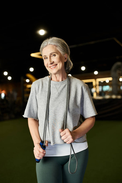 deportista madura positiva con el pelo gris posando con saltar la cuerda y sonriendo a la cámara en el gimnasio - Foto, imagen