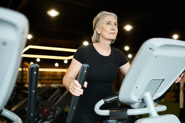 good looking jolly mature sportswoman in black t shirt exercising actively on elliptical trainer - Photo, Image