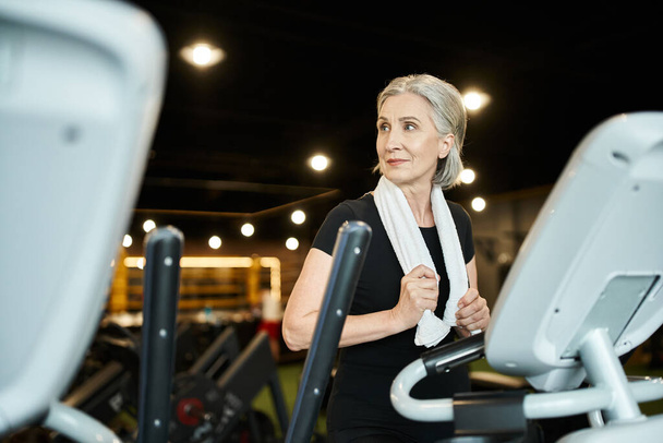jolly athletic senior woman exercising on cross trainer while in gym with towel on shoulders - Photo, Image