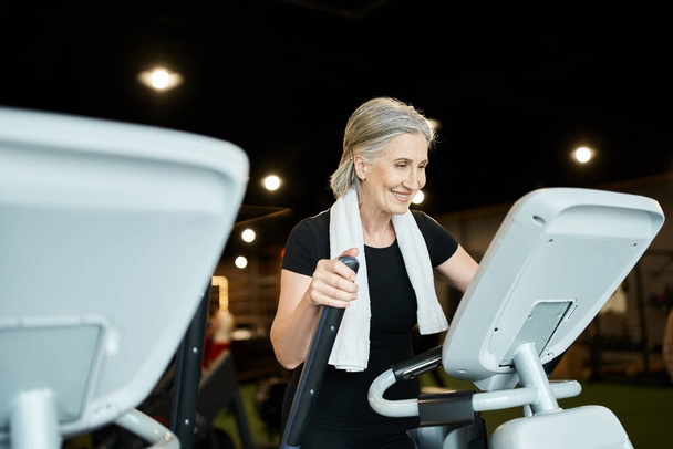 mujer alegre madura deportiva con pelo gris y toalla en los hombros haciendo ejercicio en cross trainer - Foto, imagen