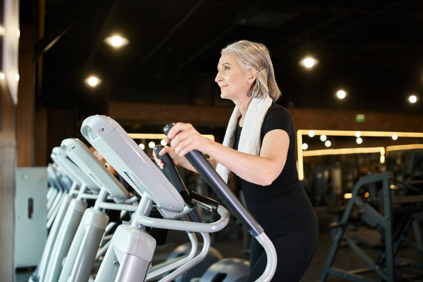 appealing senior cheerful sportswoman with towel on shoulders exercising on elliptical trainer - Photo, Image