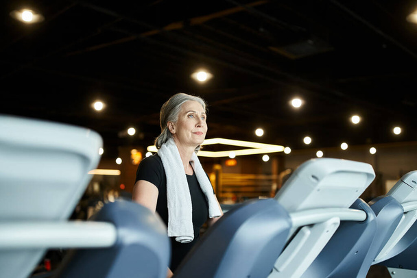 appealing mature sportswoman in black t shirt with towel on shoulders training on treadmill actively - Photo, Image