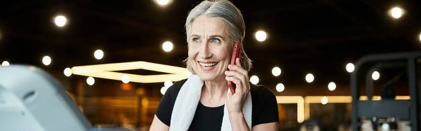 mujer mayor positiva con toalla en los hombros hablando por teléfono, mientras que en la cinta de correr en el gimnasio, pancarta - Foto, Imagen