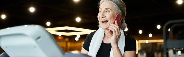 mulher madura alegre com toalha nos ombros falando por telefone enquanto na esteira no ginásio, banner - Foto, Imagem
