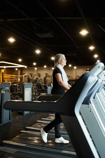 good looking mature sporty woman in black attire training on treadmill actively while in gym - Foto, afbeelding