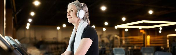 ηλικιωμένη χαρούμενη γυναίκα με γκρίζα μαλλιά και ακουστικά που γυμνάζονται στο διάδρομο γυμναστικής, banner - Φωτογραφία, εικόνα