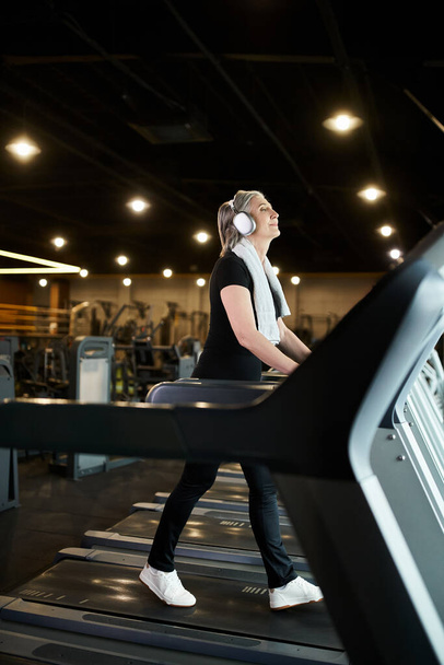 mujer alegre madura de buen aspecto con pelo gris y auriculares haciendo ejercicio en la cinta de correr en el gimnasio - Foto, imagen