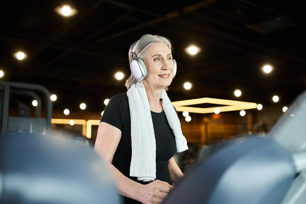 dobrze wyglądająca starsza, wesoła kobieta z siwymi włosami i słuchawkami ćwicząca na bieżni w siłowni - Zdjęcie, obraz