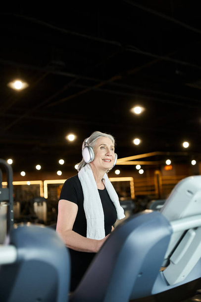 joyful pretty senior woman in cozy attire training on treadmill and listening music in headphones - Photo, Image