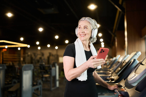 alegre mujer mayor con toalla y auriculares sosteniendo el teléfono y haciendo ejercicio en la cinta activamente - Foto, imagen