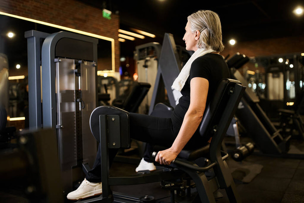 atractiva mujer mayor de pelo gris en traje deportivo ejercitando activamente en la máquina de becerro en el gimnasio - Foto, Imagen