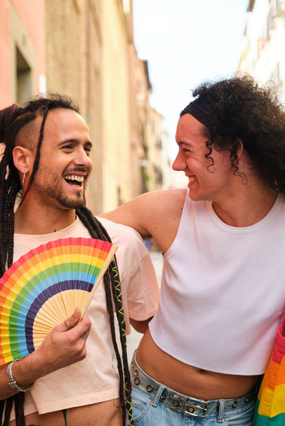 Couple gay riant et souriant tout en marchant dans une rue pendant la parade de la fierté. L'un d'eux a un ventilateur de drapeau arc-en-ciel. - Photo, image