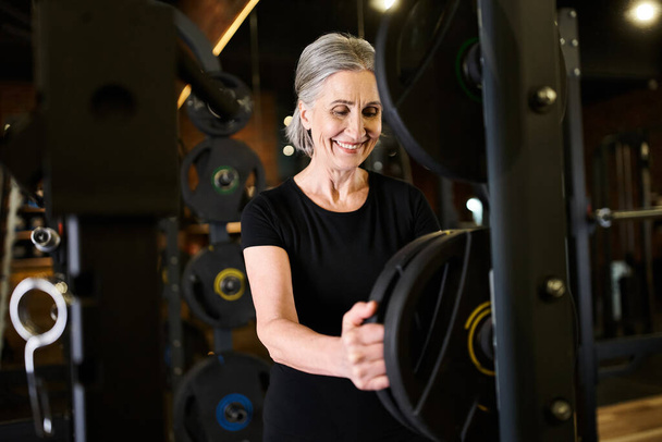 mujer senior de pelo gris positivo en ropa deportiva mirando hacia otro lado junto a los discos de peso mientras está en el gimnasio - Foto, imagen