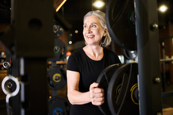 χαρούμενη γκρίζα μαλλιά ηλικιωμένη γυναίκα σε ζεστή ενδυμασία κοιτάζοντας μακριά δίπλα σε δίσκους βάρους, ενώ στο γυμναστήριο - Φωτογραφία, εικόνα
