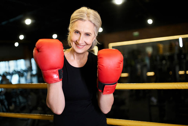довольно веселая спортивная старшая женщина в боксёрских перчатках, улыбающаяся в камеру во время выступления на ринге в спортзале - Фото, изображение