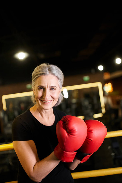 good looking cheerful senior sportswoman with boxing gloves smiling at camera while on ring in gym - Photo, Image