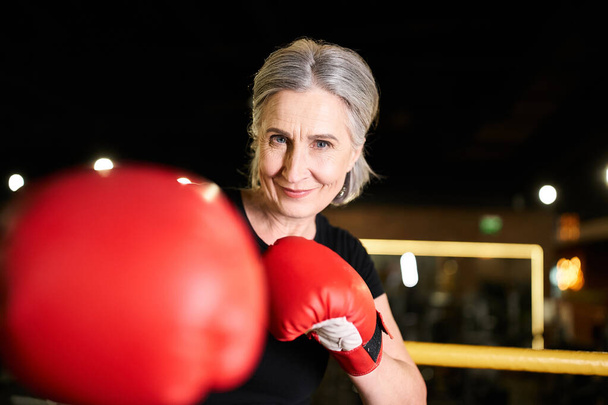 довольно позитивная старшая спортсменка с боксерскими перчатками тренирующаяся на ринге и улыбающаяся перед камерой в тренажерном зале - Фото, изображение