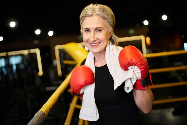 good looking joyful mature sportswoman with towel and boxing gloves smiling at camera on ring - Photo, Image