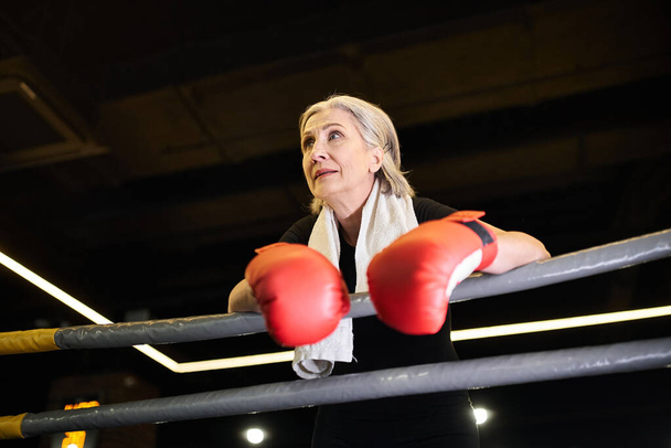 sportowe siwe włosy dojrzała kobieta z ręcznikiem i rękawice bokserskie patrząc daleko podczas gdy na ringu w siłowni - Zdjęcie, obraz