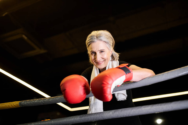 vrolijke atletische volwassen vrouw met handdoek en bokshandschoenen oefenen op ring en glimlachen op de camera - Foto, afbeelding