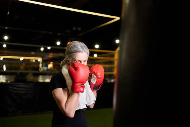 mujer madura de buen aspecto con pelo gris y toalla en guantes de boxeo golpeando saco de boxeo en el gimnasio - Foto, imagen
