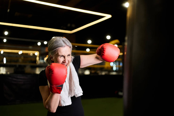αθλητική όμορφη ηλικιωμένη γυναίκα με γκρίζα μαλλιά σε γάντια πυγμαχίας ξυλοδαρμό σάκο του μποξ στο γυμναστήριο - Φωτογραφία, εικόνα
