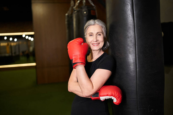 mature athletic jolly sportswoman posing with boxing gloves near punching bag and smiling at camera - Photo, Image