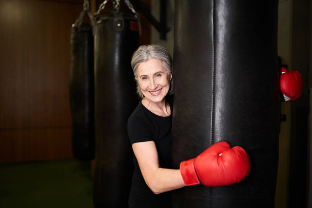 radosna dojrzała kobieta w odzieży sportowej w rękawiczkach bokserskich w pobliżu worka bokserskiego i uśmiechnięta do kamery - Zdjęcie, obraz