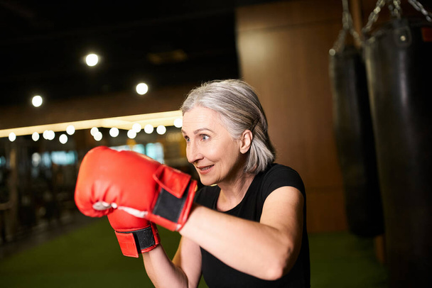 atlética madura atractiva mujer con pelo gris posando con guantes de boxeo mientras entrena en el gimnasio - Foto, imagen