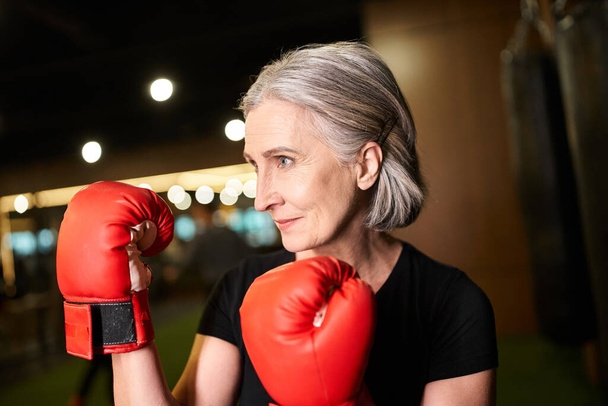 sportovní dobře vypadající starší žena s šedými vlasy pózovat s boxerskými rukavicemi při tréninku v tělocvičně - Fotografie, Obrázek